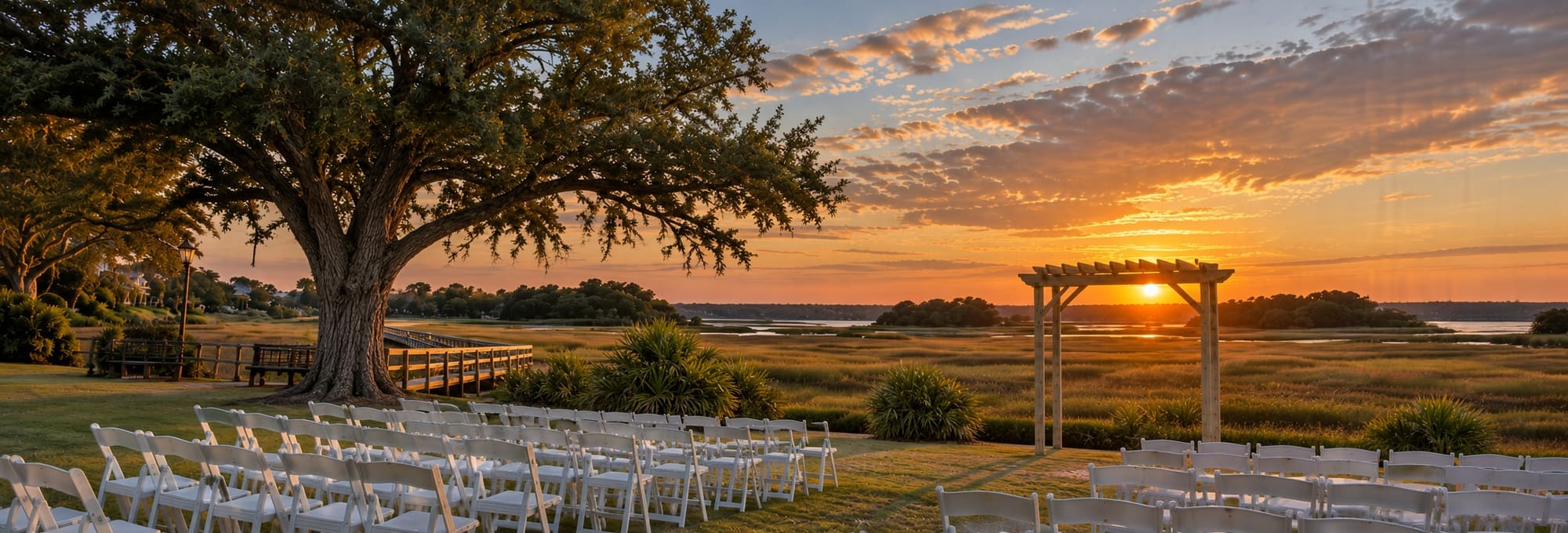 Ceremony chairs and a wooden arbor facing the marsh and river at sunset, beneath a live oak