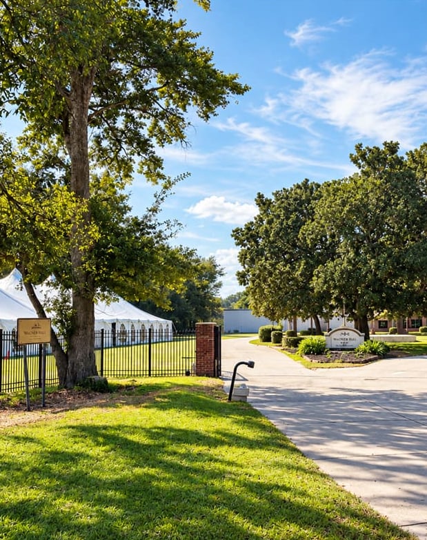The Wagner Hall entrance drive and sign beneath live oaks, with a white tent beyond the fence