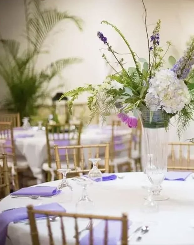 A reception table set with white linens, lavender napkins and a tall floral centerpiece